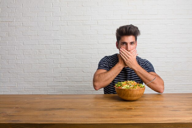 Hombre joven hermoso y natural sentado en una mesa que cubre la boca