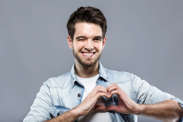 El hombre joven hermoso que hace un corazón con el suyo entrega el fondo gris.