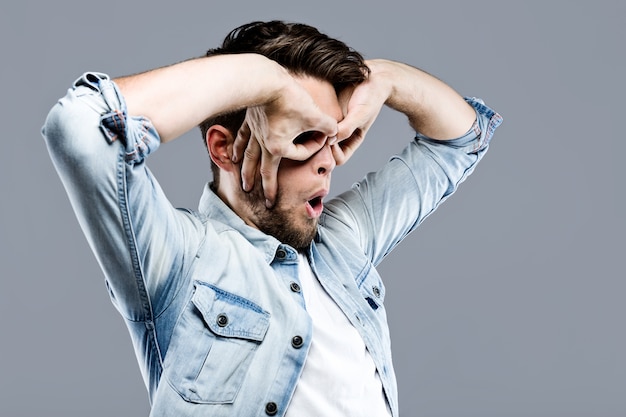 Hombre joven hermoso que hace los vidrios del dedo sobre fondo gris.