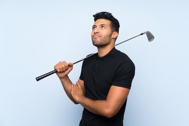 Hombre joven hermoso que juega a golf sobre la pared azul aislada que hace gesto de las dudas mientras que levanta los hombros