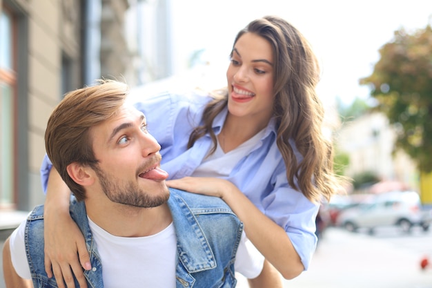 Hombre joven hermoso que lleva a mujer joven atractiva sobre los hombros mientras pasan tiempo juntos al aire libre.