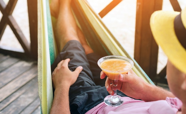 Hombre joven hermoso que miente en una hamaca en una playa soleada por un océano y sostenga el cóctel en manos. El sol brilla brillante.