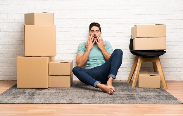 Hombre joven hermoso que se mueve en nuevo hogar entre las cajas con la expresión facial de la sorpresa