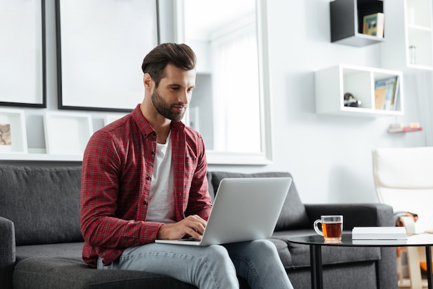 Hombre joven hermoso que se sienta en el sofá usando la computadora portátil.