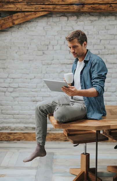 Hombre joven hermoso que usa la tableta digital y srinking el café mientras que se sienta en el cuarto rústico