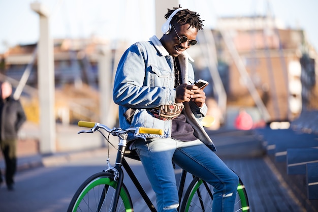 Hombre joven hermoso que usa el teléfono móvil y la bicicleta fija del engranaje en la calle.