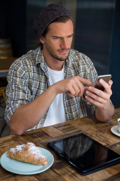 Hombre joven hermoso que usa el teléfono móvil en la mesa en la cafetería