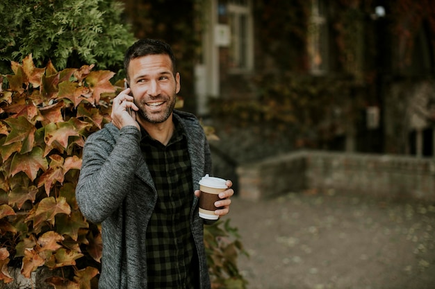 Hombre joven hermoso que usa el teléfono móvil y que sostiene el café para llevar en el parque
