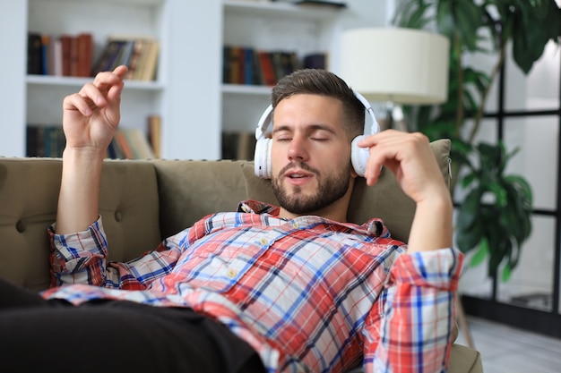 Hombre joven inconformista se acuesta en el sofá y escucha música con auriculares inalámbricos.