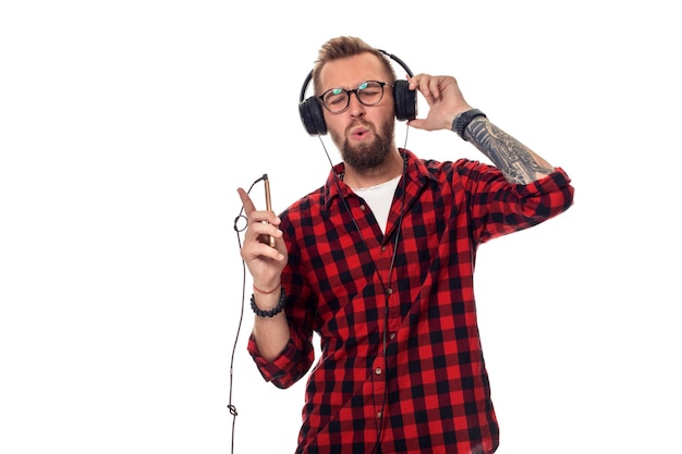 Hombre joven inconformista en camisa a cuadros y gafas con auriculares mirando feliz sobre fondo blanco.