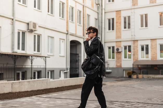 Hombre joven inconformista en gafas de sol de moda con un elegante desgaste con una mochila posando y fuma en la calle cerca de edificios antiguos. El modelo de chico moderno con ropa de gran tamaño disfruta de fumar. Receso para fumar