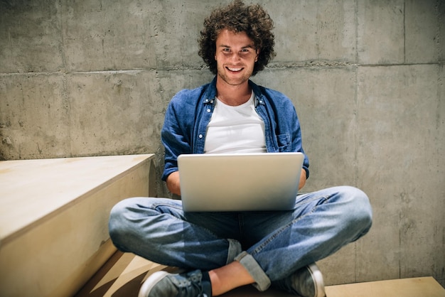 Hombre joven inteligente con cabello rizado usando una computadora portátil para trabajar navegando en línea en Internet sentado en las escaleras en el muro de hormigón Hombre guapo leyendo y escribiendo en su computadora portátil en la oficina