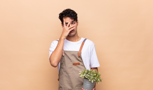 Hombre joven jardinero que parece sorprendido, asustado o aterrorizado, cubriendo la cara con la mano y mirando entre los dedos