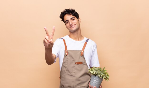 Hombre joven jardinero sonriendo y mirando feliz, despreocupado y positivo, gesticulando victoria o paz con una mano