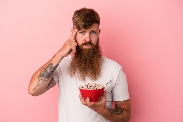 Hombre joven de jengibre caucásico con barba larga sosteniendo un cuenco de cereales aislado sobre fondo rosa señalando la sien con el dedo, pensando, centrado en una tarea.