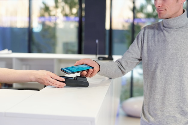 Hombre joven en jersey gris con teléfono inteligente con página de banca en línea en la pantalla durante el proceso de pago sin contacto en la tienda o centro de ocio