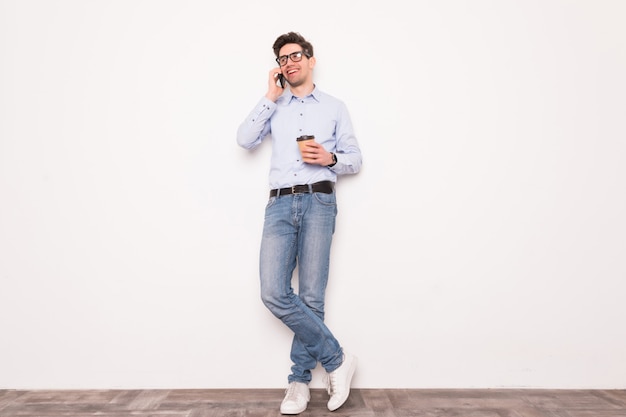 Hombre joven joven que habla en el teléfono con la taza de café aislada en el espacio blanco. Retrato de hombre seguro, estilo de vida moderno, concepto de comunicación