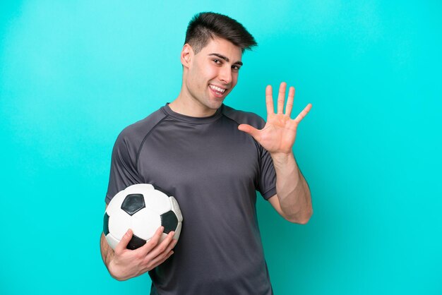 Hombre joven jugador de fútbol caucásico aislado sobre fondo azul contando cinco con los dedos