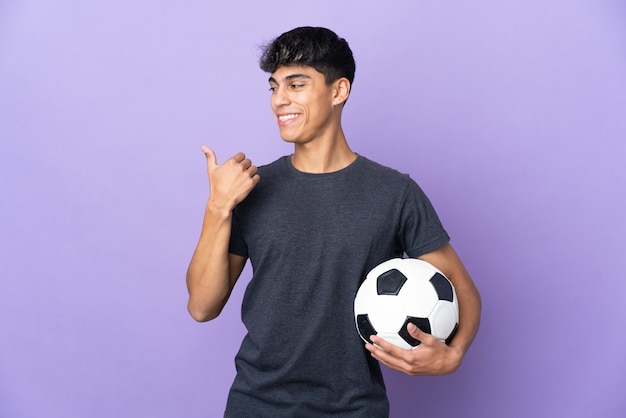 Hombre joven jugador de fútbol sobre fondo púrpura aislado apuntando hacia el lado para presentar un producto