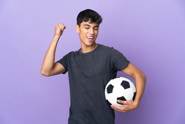 Hombre joven jugador de fútbol sobre pared púrpura aislada celebrando una victoria