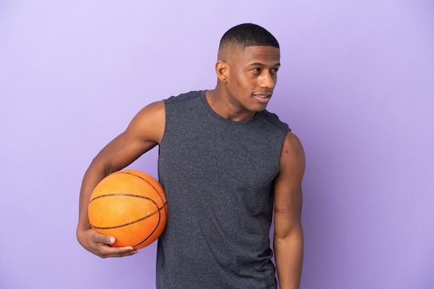 Hombre joven jugador latino de baloncesto aislado en púrpura mirando hacia el lado y sonriendo