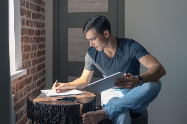 Hombre joven lindo en una camiseta y pantalones vaqueros en una mesa de madera con una tableta, escribe en un cuaderno, plan de negocios, independiente, inicio.