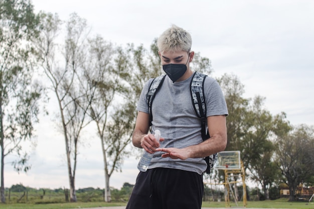 Hombre joven con una máscara aplicando spray de alcohol en sus manos en un campo abandonado.