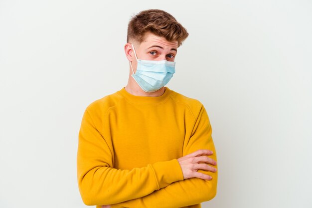 Hombre joven con una máscara para el coronavirus aislado sobre fondo blanco infeliz mirando a puerta cerrada con expresión sarcástica.