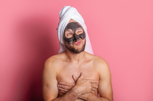 Hombre joven con una máscara cosmética en su rostro, guiña pensativamente con un ojo sobre un fondo rosa.