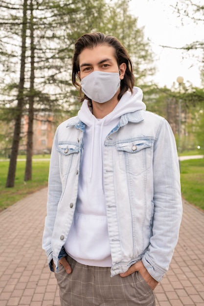 Hombre joven con máscara protectora, sudadera con capucha blanca y chaqueta de mezclilla ligera manteniendo las manos en los bolsillos de los pantalones