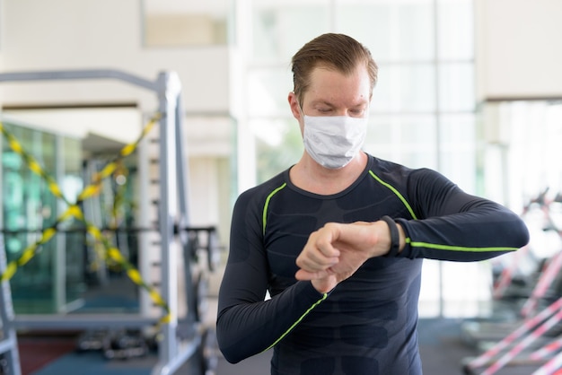 Hombre joven con máscara para protegerse del brote de coronavirus comprobando el reloj inteligente en el gimnasio durante el coronavirus covid-19