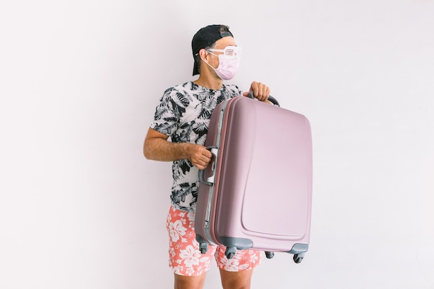 Hombre joven con una máscara para protegerse del covid-19 de vacaciones yendo de viaje, vestido con una camisa floral y gorra, sosteniendo una maleta, a la luz del día en una pared blanca