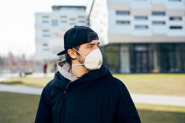 Hombre joven con máscara respiratoria en la calle y mirando a un lado, hombre de pie al aire libre con ropa casual y protector facial durante la pandemia
