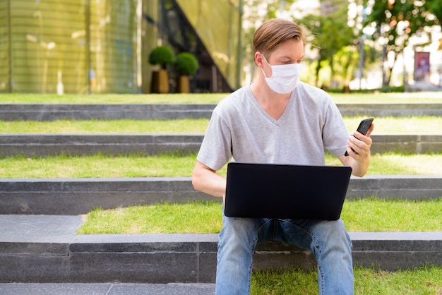 Hombre joven con máscara con teléfono y portátil mientras está sentado al aire libre