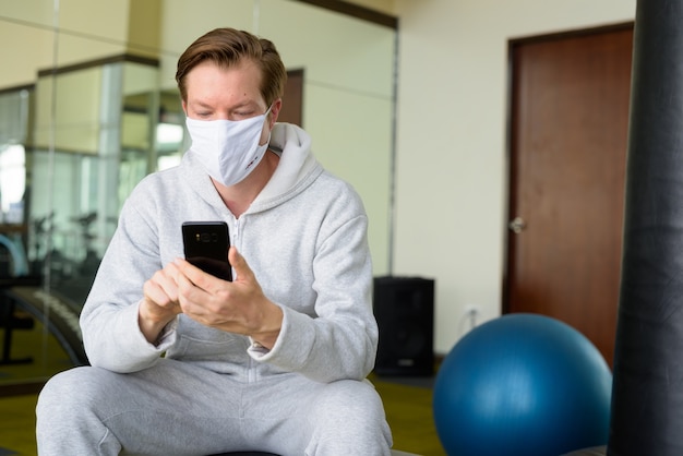 Hombre joven con máscara usando teléfono y sentado en el gimnasio