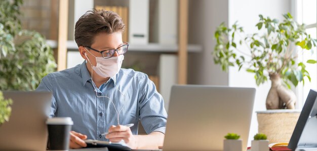 Hombre joven en mascarilla está trabajando en una computadora portátil en la oficina.