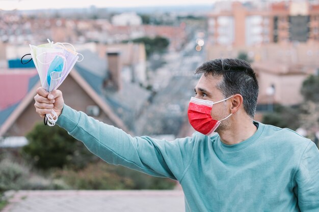 Hombre joven con una mascarilla quirúrgica roja sosteniendo un puñado de mascarillas quirúrgicas de colores con la mano. Concepto Covid-19