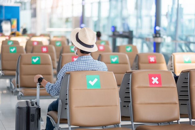 Foto hombre joven con mascarilla sentado en una silla en la terminal del aeropuerto, protección contra la enfermedad del coronavirus (covid-19), viajero hipster listo para viajar. nuevos conceptos de distanciamiento normal y social