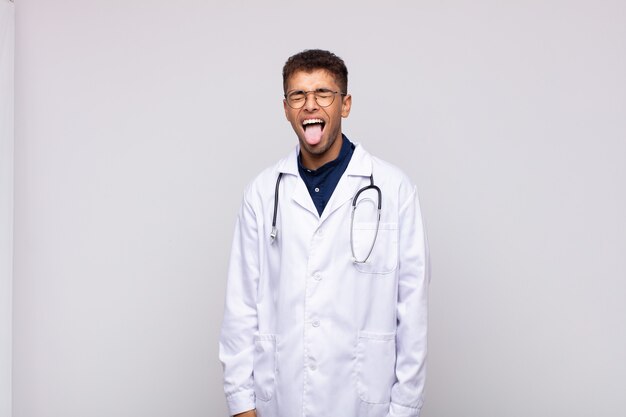 Hombre joven médico con actitud alegre, despreocupada y rebelde, bromeando y sacando la lengua, divirtiéndose