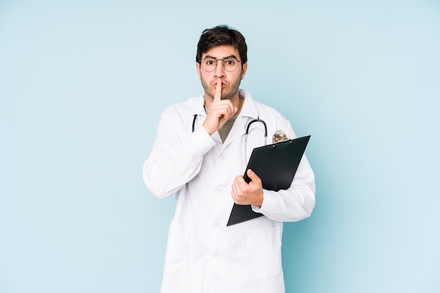 Hombre joven médico aislado sobre fondo azul manteniendo un secreto o pidiendo silencio.