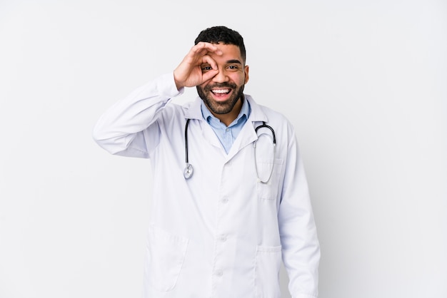 Hombre joven médico árabe aislado emocionado manteniendo el gesto ok en el ojo.