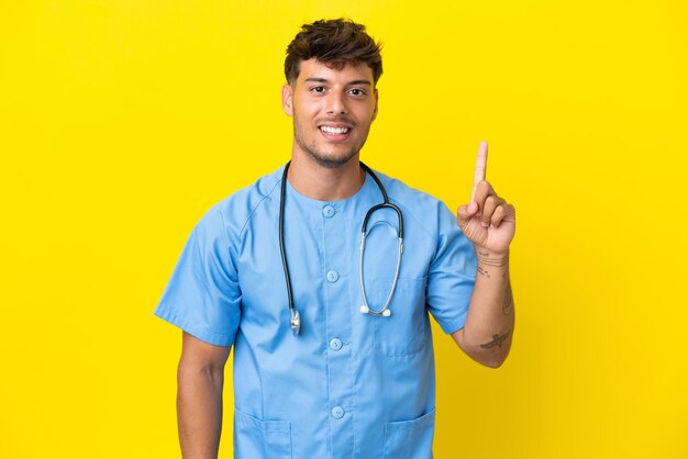 Hombre joven médico cirujano aislado sobre fondo amarillo mostrando y levantando un dedo en señal de lo mejor