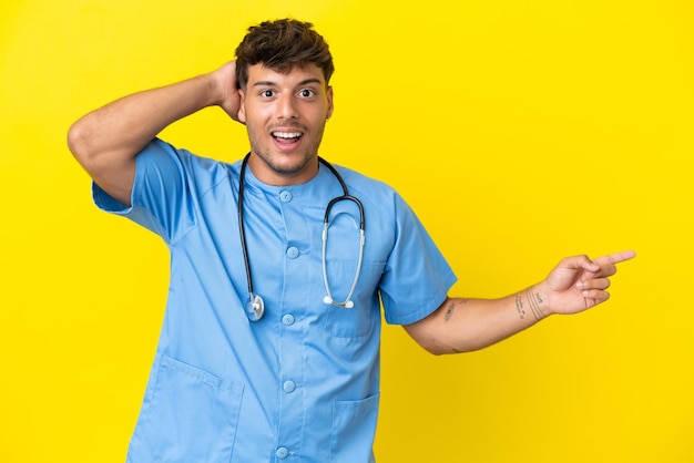 Hombre joven médico cirujano aislado sobre fondo amarillo sorprendido y apuntando con el dedo hacia el lado