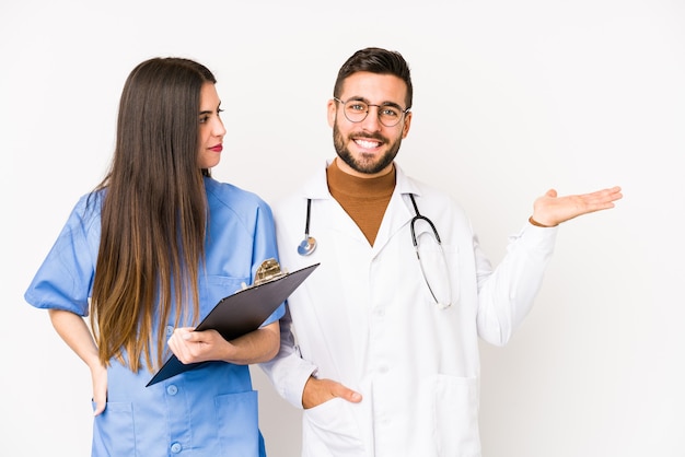 Hombre joven médico y una enfermera aislados mostrando un espacio de copia en una palma y sosteniendo otra mano en la cintura.