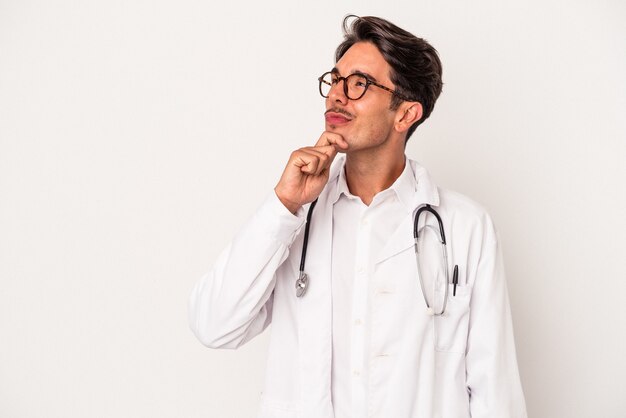 Hombre joven médico de raza mixta aislado sobre fondo blanco mirando hacia los lados con expresión dudosa y escéptica.