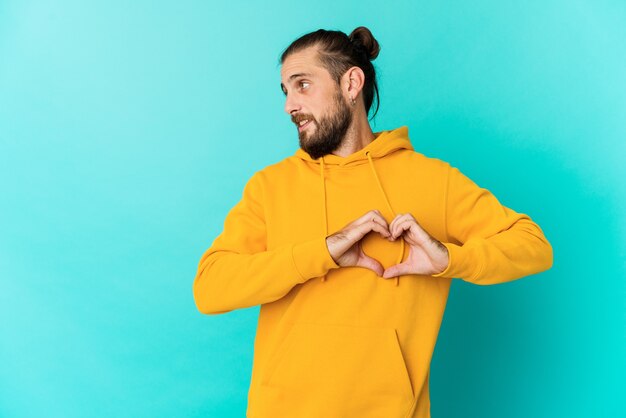 Hombre joven con mirada de pelo largo sonriendo y mostrando una forma de corazón con las manos.