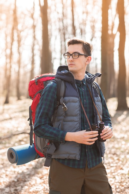 Hombre joven con mochila y con un binocular en sus manos, caminando en el bosque