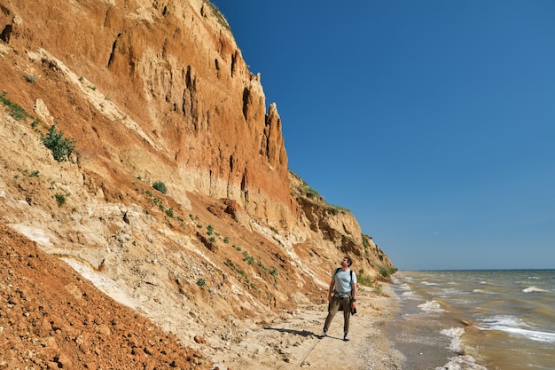 Hombre joven y mochila camina solo a lo largo de una colina de arena sobre el mar.