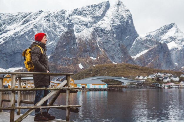 Hombre joven con una mochila de pie sobre un muelle de madera el fondo de montañas nevadas y lago