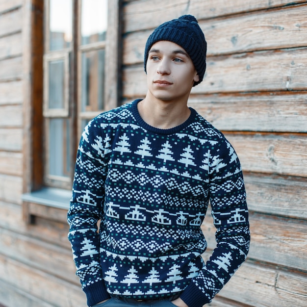 Hombre joven de moda con un sombrero de moda vintage en un elegante suéter de año nuevo de punto de invierno azul está posando cerca de una casa de pueblo de madera al aire libre. Chico moderno con estilo.